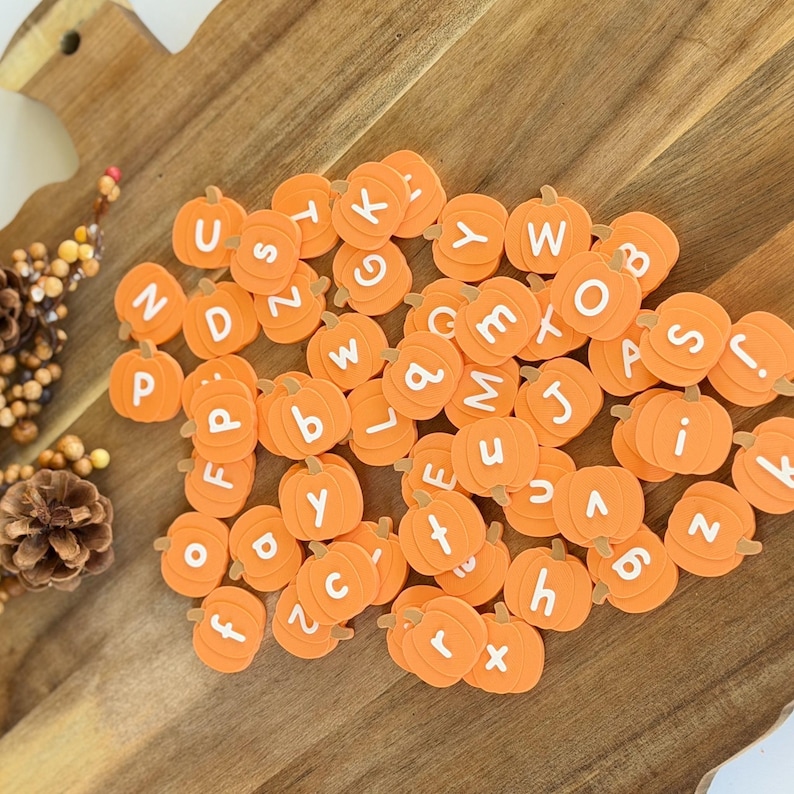 May include: A collection of small, orange, pumpkin-shaped beads, each featuring a single white letter printed on the front. The pumpkin beads are scattered across a wooden surface, displaying a variety of uppercase and lowercase alphabet letters.