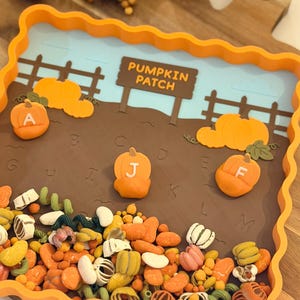 May include: A square orange tray with a scalloped edge featuring a pumpkin patch scene. The tray contains a brown field with letters A through M, a sign reading PUMPKIN PATCH, and three orange pumpkin figures labeled A, J, and F.