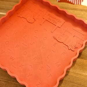 May include: A square, orange silicone mold with a scalloped edge featuring an embossed alphabet from A to Z, wavy lines, two pumpkin illustrations, and a sign that reads PUMPKIN PATCH. The mold has a flat base and raised sides for casting.
