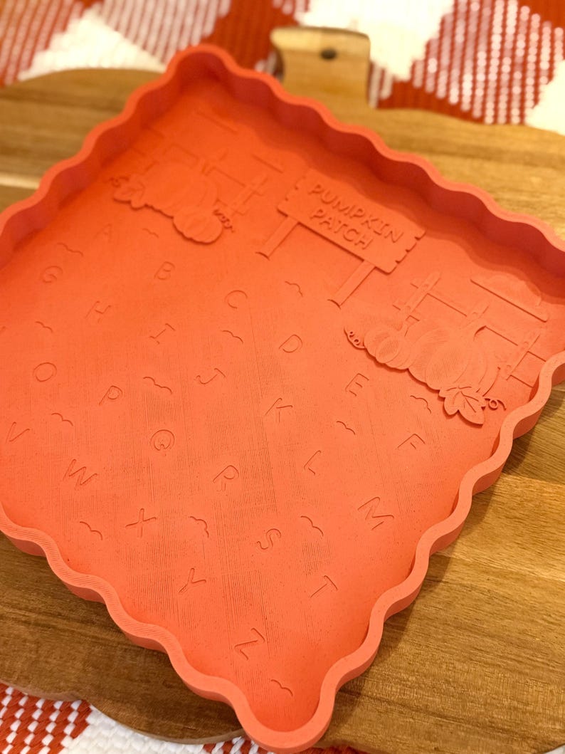 May include: A square, orange silicone mold with a scalloped edge featuring an embossed alphabet from A to Z, wavy lines, two pumpkin illustrations, and a sign that reads PUMPKIN PATCH. The mold has a flat base and raised sides for casting.
