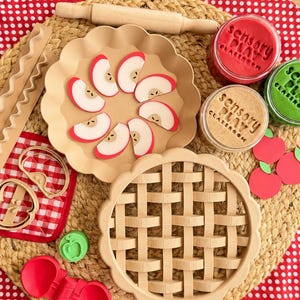 May include: A children's play set featuring a beige plastic pie dish with apple slices, a lattice pie crust top, a wooden rolling pin, two plastic apple cutters, a red folding tool, and three jars of sensory play dough labeled Sensory Play Classroom.