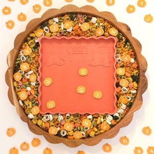 May include: A round wooden scalloped tray filled with multicolored dried pasta and small orange pumpkin-shaped tokens featuring letters and numbers. A square orange silicone mat with a pumpkin patch design and embossed alphabet letters sits in the center.