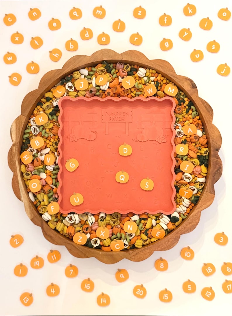 May include: A round wooden scalloped tray filled with multicolored dried pasta and small orange pumpkin-shaped tokens featuring letters and numbers. A square orange silicone mat with a pumpkin patch design and embossed alphabet letters sits in the center.