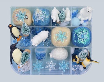 May include: A clear plastic storage container divided into twelve compartments filled with winter-themed sensory items including white polar bear figurines, black and white penguin figures, small green bottle brush trees, blue glittery slime, an igloo, and blue snowflake accents.
