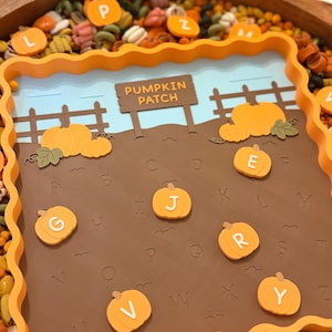May include: A square orange tray with a scalloped edge containing a brown board printed with the alphabet and a pumpkin patch scene. The scene features a brown sign reading PUMPKIN PATCH, a fence, and several orange pumpkin-shaped letter tokens.