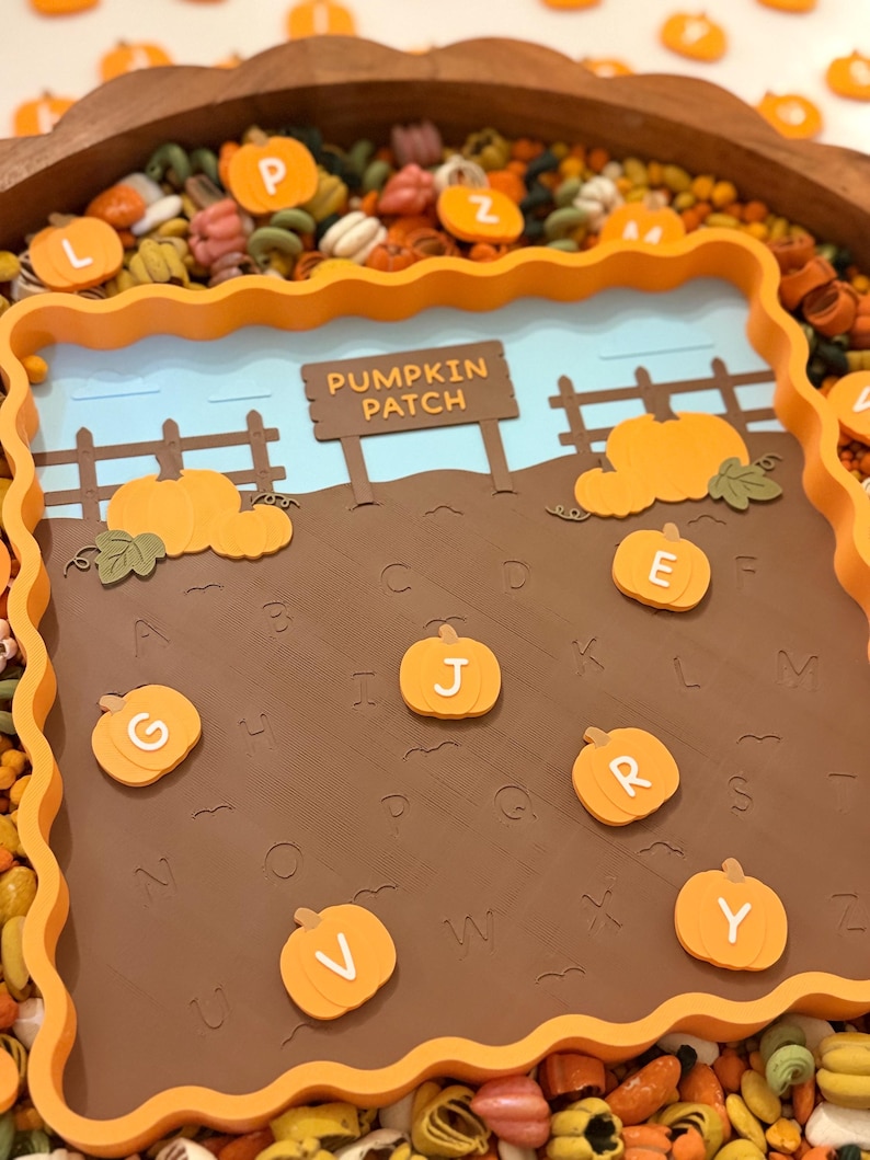 May include: A square orange tray with a scalloped edge containing a brown board printed with the alphabet and a pumpkin patch scene. The scene features a brown sign reading PUMPKIN PATCH, a fence, and several orange pumpkin-shaped letter tokens.