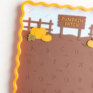 May include: A square tray with a wavy yellow border featuring a brown field with embossed letters of the alphabet from A to Y. The scene includes a brown wooden fence, a sign that reads PUMPKIN PATCH, and several orange pumpkins.