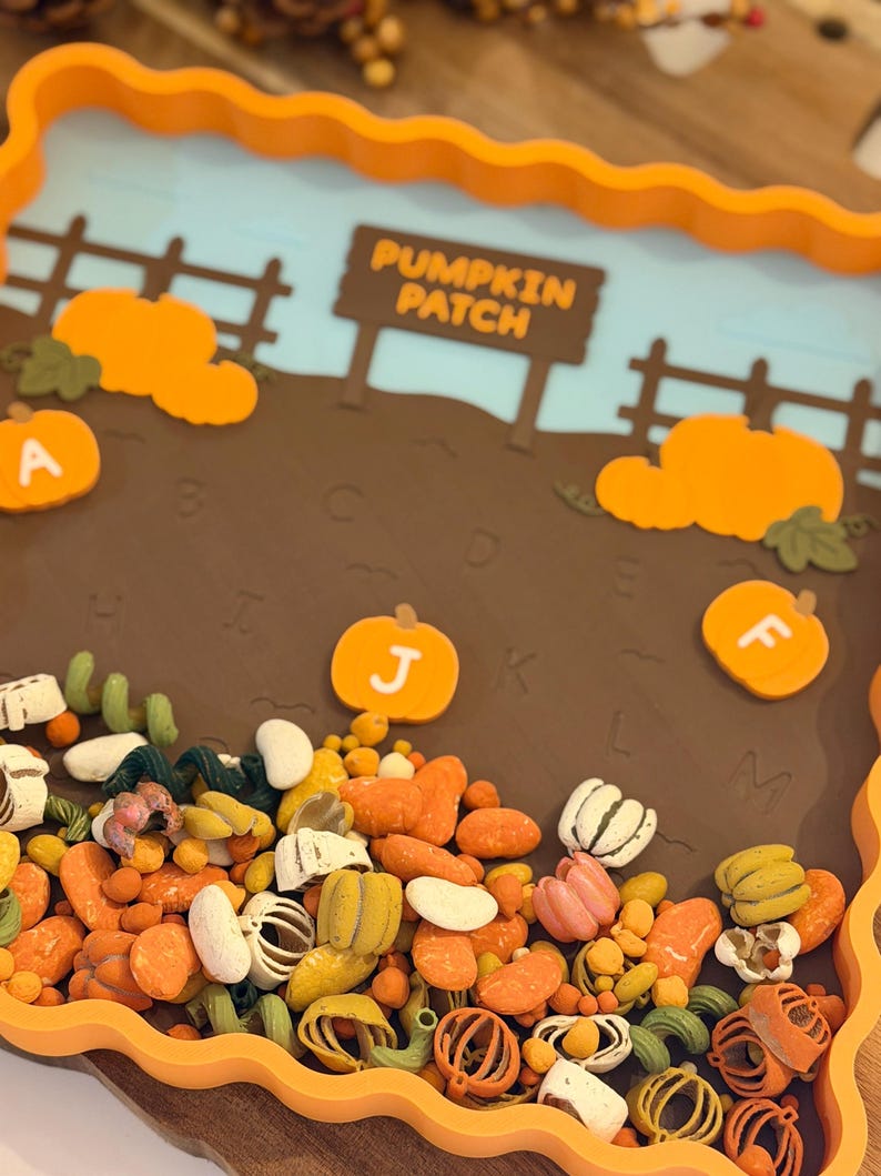 May include: A rectangular orange tray with a scalloped edge featuring a brown field background with a sign that reads PUMPKIN PATCH. The tray includes orange pumpkin cutouts labeled with letters A, J, and F, and a base filled with assorted small colorful autumn-themed sensory items like miniature pumpkins, beans, and pasta shapes.