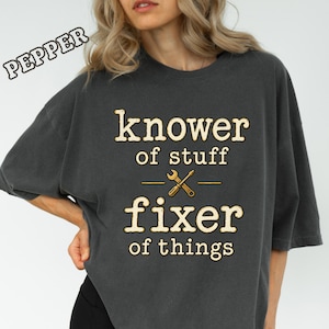 May include: A charcoal gray short-sleeve t-shirt featuring the text "knower of stuff" above a graphic of a crossed wrench and screwdriver, followed by the text "fixer of things" in a light beige font. The word "PEPPER" is printed in the top left.