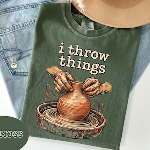 May include: A moss green t-shirt featuring a graphic of hands shaping a clay pot on a pottery wheel. The text "i throw things" is printed in a cream-colored font above the illustration. The shirt has a crew neckline and short sleeves.