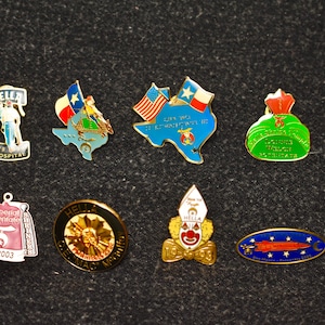 May include: A collection of colorful enamel pins with various designs. The pins include a Texas-shaped pin with flags, a clown, a hospital logo, and other emblems. The pins are on a dark gray surface.