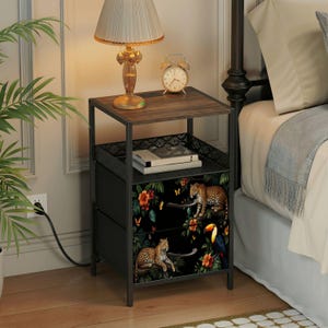 May include: A black nightstand with two drawers, a top shelf, and a metal frame. The drawer fronts feature a black background with a floral and animal print. A lamp and alarm clock sit on top.