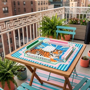 Puede incluir: Una mesa plegable de madera con un juego de Mahjong colorido. El tablero presenta un dise&ntilde;o de tigre con detalles en rosa, azul y verde. El juego incluye fichas verdes y rosas, sobre una alfombra a rayas azules y rosas.