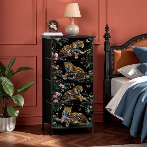 May include: A black four-drawer chest with a leopard print design. Each drawer features a leopard resting on a branch, surrounded by pink flowers and butterflies. The chest has a black top and a metal frame.