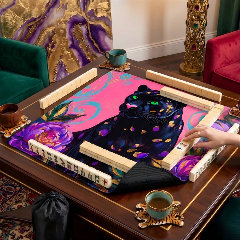 May include: A mahjong set with a pink and purple mat featuring a black panther design. The set includes tiles, a black bag, and tiger-print coasters. The table is made of dark wood.