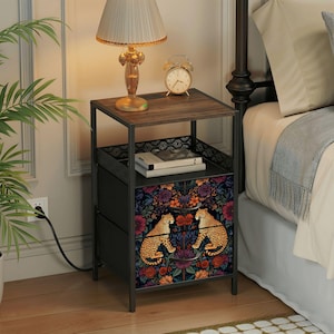 May include: A black metal bedside table with a dark wood top, a floral patterned drawer, and two drawers. The floral pattern features two cheetahs and colorful flowers. A gold lamp and alarm clock sit on top.