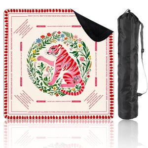 Peut inclure: Un tapis de jeu carr&eacute; avec une illustration de tigre rose entour&eacute;e d'une couronne florale. Le tapis a une bordure rouge avec du texte et est accompagn&eacute; d'un sac de transport noir. Le tapis est con&ccedil;u pour un jeu.