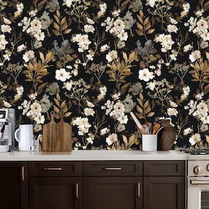 May include: Black floral wallpaper featuring a repeating pattern of white and gold flowers and leaves. The wallpaper is installed on a wall behind a kitchen countertop with dark brown cabinets and a white countertop. Kitchen accessories are visible.