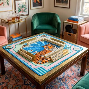 Puede incluir: Una mesa de centro cuadrada de madera con un marco de metal color lat&oacute;n. La mesa presenta un tablero de juego de mahjong colorido con un dise&ntilde;o de tigre. Las piezas del juego est&aacute;n dispuestas en el tablero. La habitaci&oacute;n tiene un sill&oacute;n verde de terciopelo y sillas rosas.