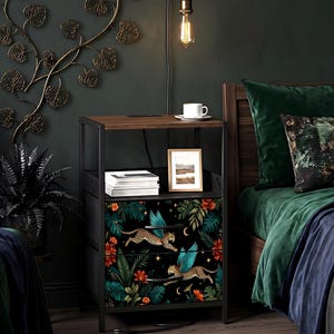 May include: A black nightstand with a dark wood top and metal frame. The drawers feature a vibrant design of flying leopards with blue wings, set against a backdrop of lush foliage and stars. A framed picture and books sit on the shelf.