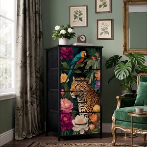 May include: A black chest of drawers with a vibrant floral and animal design. The drawers feature a parrot, leopard, and colorful flowers. The chest is in a room with green walls, a window, and a green velvet chair.