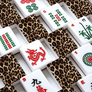 May include: A set of white Mahjong tiles with red, green, and black symbols and numbers. The tiles are arranged in a grid, interspersed with leopard print patterned dividers. The game pieces are in a white box.
