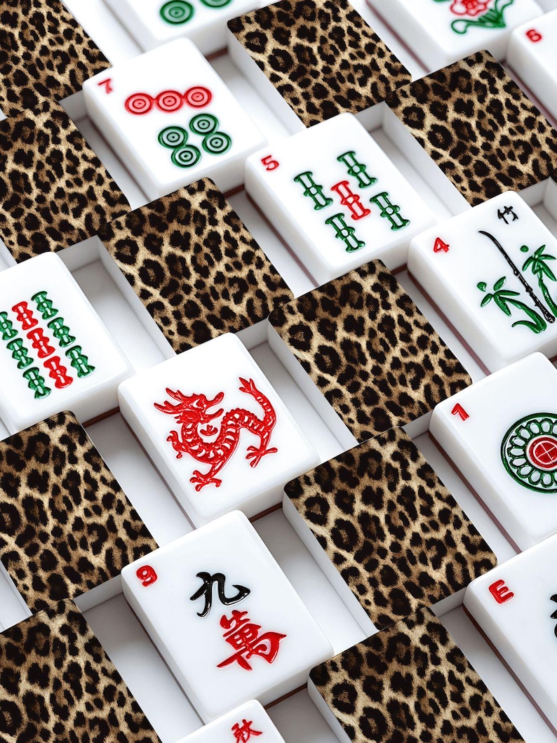 Puede incluir: Un juego de fichas de Mahjong blancas con s&iacute;mbolos y n&uacute;meros rojos, verdes y negros. Las fichas est&aacute;n dispuestas en una cuadr&iacute;cula, intercaladas con divisores con estampado de leopardo. Las piezas del juego est&aacute;n en una caja blanca.