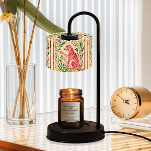 May include: A black candle warmer with a cylindrical lampshade featuring a pink tiger design amidst floral patterns. A lit candle sits below. The lamp has a curved arm and a round base.