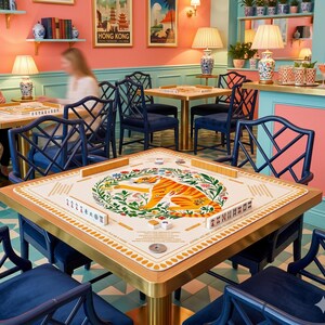 Peut inclure: Une table de mahjong carr&eacute;e avec un cadre dor&eacute; et une surface de jeu cr&egrave;me. La surface pr&eacute;sente une illustration de tigre entour&eacute;e de motifs floraux. Des chaises bleu fonc&eacute; entourent la table, et la pi&egrave;ce a une esth&eacute;tique r&eacute;tro.