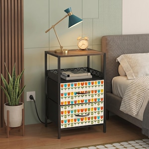 May include: A bedside table with a dark wood top, a black metal frame, and two fabric drawers featuring a repeating pattern of orange, yellow, and blue tulips on a cream background. The table includes a shelf and two black curved drawer handles.