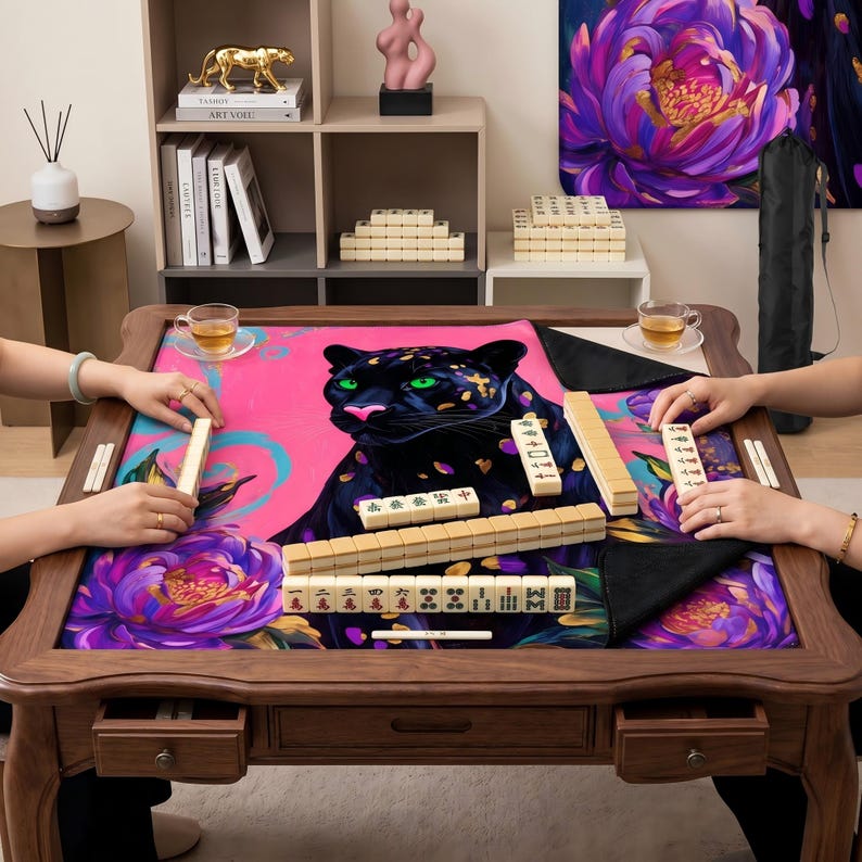 May include: A wooden mahjong table featuring a pink and purple floral design and a black panther illustration. The table has drawers and is set up for a game, with tiles and tea cups visible. A game of mahjong is in progress.