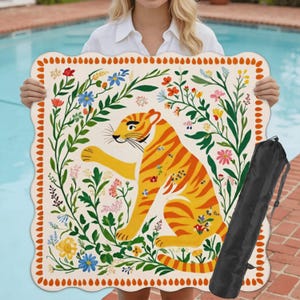May include: A cream-colored beach mat with a scalloped edge, featuring a vibrant illustration of a tiger surrounded by colorful flowers and foliage. Includes a black carrying bag. The mat is held up by a person.