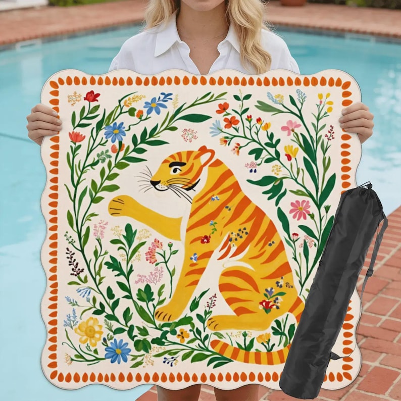 May include: A cream-colored beach mat with a scalloped edge, featuring a vibrant illustration of a tiger surrounded by colorful flowers and foliage. Includes a black carrying bag. The mat is held up by a person.