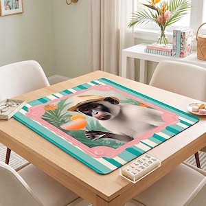 May include: A square mousepad featuring a monkey wearing a straw hat and holding a cocktail. The image is framed with a pink border and a teal and white striped background. The mousepad is on a wooden table.