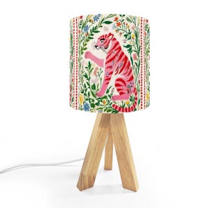 May include: A decorative table lamp with a floral design and a pink tiger illustration. The lamp has a wooden tripod base and a white cord. The lampshade features a colorful design of flowers and a pink tiger.