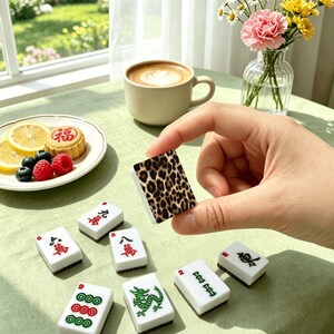 Puede incluir: Una colecci&oacute;n de fichas de Mahjong blancas y verdes con s&iacute;mbolos rojos y verdes, y una ficha con estampado de leopardo. Las fichas est&aacute;n dispuestas sobre una superficie verde claro, con una taza de caf&eacute; y un plato de fruta en el fondo.