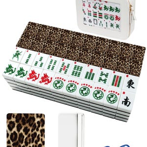Puede incluir: Un juego de Mahjong blanco con una alfombra con estampado de leopardo y un estuche a juego. El juego incluye fichas blancas con s&iacute;mbolos verdes, rojos y negros, dos dados azules y un soporte blanco para fichas.
