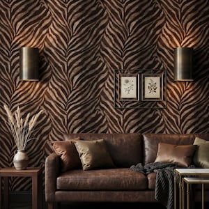 Puede incluir: Interior de salón con un sofá de cuero marrón y cojines contra un fondo de papel pintado con estampado de cebra. Dos apliques de pared iluminan el espacio. También se ven grabados botánicos enmarcados y una mesa auxiliar con un jarrón.