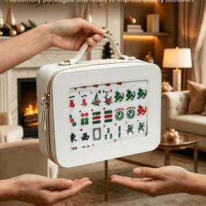 Może przedstawiać: Biały zestaw do gry w Mahjong w kwadratowej walizce z uchwytem i złotym zamkiem błyskawicznym. Zestaw zawiera kafelki z czerwonymi, zielonymi i czarnymi symbolami. Tekst na walizce brzmi "A Gift of Elegance".