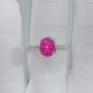 May include: A sterling silver ring featuring a vibrant pink star ruby gemstone. The oval-cut ruby is the focal point, with a star-like pattern. The band is adorned with small, sparkling accent stones.