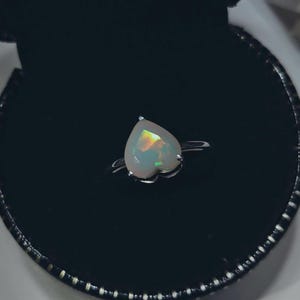 May include: A heart-shaped opal ring with a silver band. The opal has a milky white base with flashes of green, orange, and yellow. The ring is set in a black jewelry box.