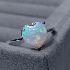 May include: A heart-shaped opal ring with a silver band. The opal has a white base with flashes of green, yellow, and orange. The ring is set on a gray surface.