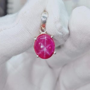 May include: A pendant with a vivid, oval star ruby gemstone. The ruby is a deep pink hue, displaying a six-ray star effect. The setting is silver-toned, with four prongs holding the stone. The pendant has a simple bail for a chain.