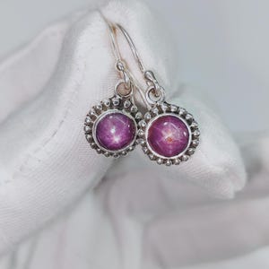 Op de afbeelding: Een paar zilveren oorbellen met ronde, bezel-set ster-robijn edelstenen. De oorbellen hebben een kralenrand en een haaksluiting. De edelstenen zijn diep roze van kleur met een zesstralig sterreneffect.