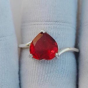 May include: A silver-toned ring featuring a prominent heart-shaped red gemstone set in a three-prong mounting. The band has a smooth, polished finish and a bypass design that curves around the central stone, creating a clean and simple aesthetic.