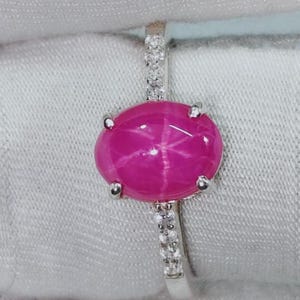 May include: A silver ring featuring a large, oval-shaped, vibrant pink star gemstone set in a four-prong mounting. The band is crafted from silver and is accented with small, clear, sparkling stones embedded along the shank on both sides.