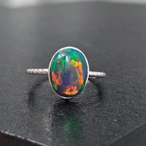 May include: A silver ring featuring a large, oval-shaped black opal gemstone. The stone displays vibrant flashes of green, orange, and red color play. The ring band has a textured, segmented design that encircles the finger.