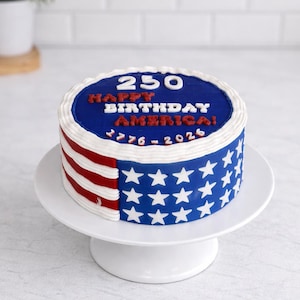 May include: A round cake decorated with the American flag design. The top is blue with white text that reads "250 HAPPY BIRTHDAY AMERICA! 1776-2026". The side features red and white stripes and white stars on a blue background.