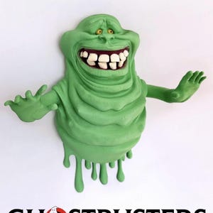 May include: A green, cartoon-like character with a wide grin, large white teeth, and yellow eyes, similar to the Ghostbusters' Slimer. The character has outstretched arms and a dripping effect. The Ghostbusters logo is at the bottom.