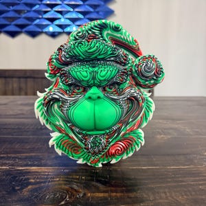 Puede incluir: Una cabeza esculpida en tres dimensiones de un personaje con gorro de Papá Noel, con un rostro verde vibrante y patrones intrincados y arremolinados en rojo, blanco, negro y verde en el pelaje y el gorro, creando un aspecto texturizado.